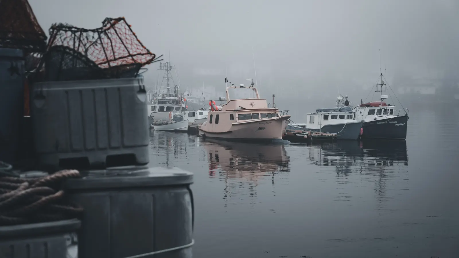 Fishing boats moored in a fog-shrouded working harbor at dawn, with traps and bins stacked on the wharf