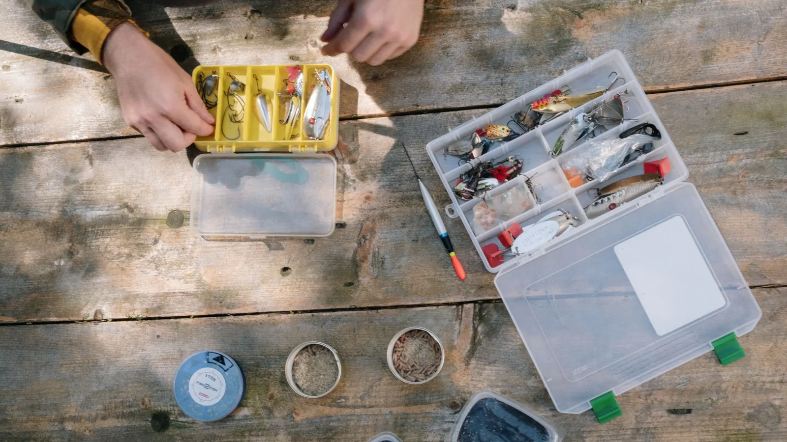 Overhead view of open tackle boxes full of lures on a weathered wood table, hands sorting through the gear