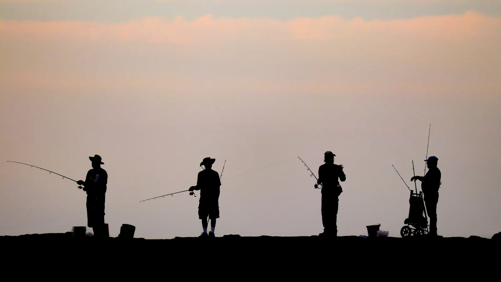 Four surf anglers silhouetted against a soft pink dawn sky, casting into the Atlantic beside their rods and bait buckets