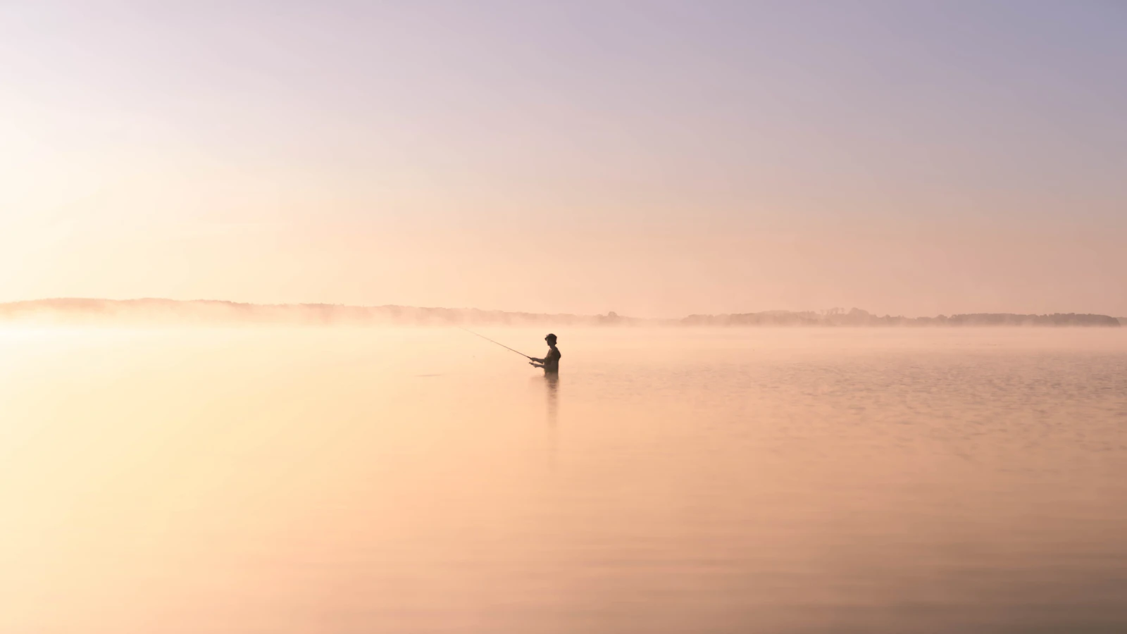A lone angler wading a mist-covered lake at sunrise, silhouetted in warm golden light