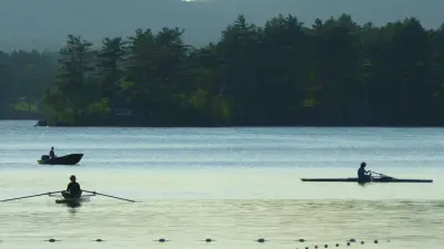 Rowers on Megunticook Lake with a wooded shore and rounded hills behind them in July 2016.